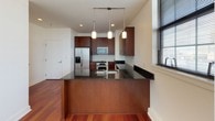 This single-floor residence offers a thoughtfully designed open-concept layout that seamlessly connects the living and kitchen areas. Rich, warm hardwood flooring flows throughout the main living spaces, complemented by crisp white walls that create a bright, clean aesthetic. The expansive living area benefits from a row of large, black-framed windows that flood the space with natural light and frame views of the surrounding urban landscape.

The kitchen is a standout feature, showcasing rich dark-wood cabinetry, sleek dark granite countertops, and a classic white subway tile backsplash. A peninsula with a matching granite surface defines the space and provides additional seating and prep area. Stainless steel appliances — including a refrigerator with water dispenser, a gas range, a built-in microwave, and a wine cooler — complete the well-equipped culinary space. Stylish pendant lighting hangs above the peninsula, adding a refined touch.

The bedrooms offer a comfortable and quiet retreat, each finished with neutral-toned carpet flooring and clean white walls. The bathroom is finished with elegant marble-look tile flooring and wall surfaces, a full soaking tub, a separate shower area with grab bars, and a vanity with ample storage — combining functionality with a refined, spa-like atmosphere throughout.