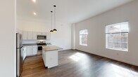 This single-floor residence offers a thoughtfully designed open-concept layout that seamlessly connects the kitchen, dining, and living areas. The kitchen is a standout feature, equipped with a full suite of stainless steel appliances — including a refrigerator, range, microwave, and dishwasher — complemented by crisp white shaker-style cabinetry, gray countertops, and a central island ideal for meal prep or casual dining. Dual pendant lights hang above the island, adding a modern, refined touch. Soaring ceilings amplify the sense of openness throughout the main living area, which is bathed in natural light from multiple large windows overlooking the surrounding urban streetscape. Rich, dark hardwood-style flooring runs consistently throughout the entire space, lending warmth and visual continuity. The bedroom is generously proportioned, with tall ceilings, additional windows for natural light, and exposed ductwork that adds an industrial character. The bathroom features the same hardwood-style flooring, a modern vanity, and direct access to a dedicated walk-in closet. Clean white walls and contemporary finishes throughout create a bright, neutral palette that is both functional and visually cohesive.