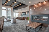 Located at 4165 Savannah Dr in De Forest, WI, this thoughtfully designed community lounge space offers an impressive blend of modern aesthetics and comfortable gathering areas across a single floor. The open-concept layout is anchored by a striking bar and kitchen island finished in warm walnut cabinetry and a crisp white quartz countertop, illuminated by elegant brass-accented glass pendant lights. A teal glass tile backsplash frames a wall-mounted flat-screen TV, adding a bold pop of color, while a herringbone hardwood floor beneath the bar area provides a refined contrast to the patterned carpet throughout the main lounge. A dedicated lounge zone features a linear fireplace set into a rich wood-paneled accent wall, flanked by channel-tufted sofas and a leather ottoman — creating a warm, inviting retreat. Floor-to-ceiling windows and glass patio doors flood the interior with natural light and frame sweeping views of the surrounding landscape. Two outdoor terraces extend the living space further, furnished with bar-height and Adirondack-style seating, ideal for taking in the open Wisconsin skyline. The overall ambiance is sophisticated yet approachable, combining contemporary design details with a welcoming, social atmosphere.