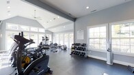 The Fitness Center at Summit at Mill Ridge is a thoughtfully designed, single-floor exercise facility built to support a well-rounded workout routine. The space features a vaulted ceiling with recessed lighting and a ceiling fan, contributing to an open, airy atmosphere. Large windows and glass-panel doors line the walls, flooding the interior with an abundance of natural light and offering views of the surrounding outdoor area. The walls are finished in a clean, neutral gray palette complemented by white trim, creating a bright and motivating environment. The rubber-floored workout area is well-organized and equipped with a comprehensive selection of fitness equipment, including cardio machines such as ellipticals, a rowing machine, and a stationary bike, as well as a weight bench and stability ball for strength and flexibility training. A dedicated free weight station features a full rack of dumbbells in a range of sizes, catering to users of all fitness levels. A posted fitness center rules board ensures the space remains welcoming and orderly for all residents. The combination of practical amenities, ample natural light, and a spacious, well-ventilated layout makes this fitness center a standout community feature at Summit at Mill Ridge.