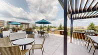 This ground-level outdoor patio is a well-appointed communal amenity space nestled within a modern multi-story building complex. The patio is defined by a clean concrete surface that extends generously to accommodate multiple distinct seating zones. A prominent pergola structure with sleek metal posts anchors one section of the space, fitted with string lights and providing partial shade over a comfortable lounge arrangement of sofas and armchairs with accent pillows, centered around a round fire feature. Adjacent to this covered zone, an open dining area features cafe-style tables and chairs, one shaded by a teal market umbrella. A built-in outdoor kitchen station with a gas grill offers a practical entertaining hub. Large decorative planters filled with vibrant seasonal foliage are positioned throughout, adding visual interest and color to the hardscape. Black metal railings define the perimeter, framing views of the surrounding lawn, mature trees, and neighboring buildings. The exterior building facade incorporates a mix of masonry block, brick, and teal-toned siding, contributing to a contemporary aesthetic. Glass doors provide direct interior access, seamlessly connecting indoor and outdoor spaces. The patio benefits from open sky exposure, ensuring abundant natural light throughout the day.