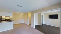 Located at 5300 New Center Dr in Wilmington, North Carolina, this single-floor space offers a clean, functional layout designed for comfortable everyday living. The main living area is generously sized, featuring warm beige walls, plush carpeting, and a ceiling fan with integrated lighting that provides both comfort and illumination. A textured ceiling adds a classic finishing touch throughout the space. Transitioning from the living area, the flooring shifts to a dark hardwood-style surface, creating a visual contrast that defines the adjoining spaces. Wide doorways and open passageways connect the rooms, contributing to an open and airy feel that promotes easy flow throughout the unit. Neutral wall tones provide a versatile backdrop that complements a variety of interior styles. HVAC vents are integrated into the ceiling, ensuring efficient climate control throughout. The layout includes what appears to be additional rooms accessible from the main living space, including areas that may serve as bedrooms or utility spaces, each benefiting from the same clean, well-maintained finishes seen throughout. Overall, the space presents a bright, neutral, and welcoming environment well-suited for a range of occupants.