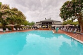 Located at 5751 Wood Meadow Way in Centreville, Virginia, this outdoor amenity space centers around a generously sized, resort-style swimming pool with a distinctive cross-shaped layout. The crystal-clear turquoise water is framed by a broad brick-paved sundeck that offers ample room for lounging and socializing. Rows of modern chaise lounge chairs are arranged along all sides of the pool, complemented by a collection of shade umbrellas that provide comfortable relief from the sun. Scattered seating areas with bistro-style tables and chairs create inviting spots for casual gatherings. The pool area is enclosed by a black wrought-iron fence, ensuring both safety and a clean aesthetic. Mature crape myrtle trees in full bloom add vibrant color and natural shade throughout the space, while manicured shrubs and ornamental plantings enhance the lush, park-like atmosphere. A two-story clubhouse building anchors one end of the pool area, featuring a covered porch and outdoor seating that overlooks the entire pool deck. The overall ambiance is relaxed and resort-inspired, offering a well-appointed outdoor retreat designed for both leisure and community enjoyment.