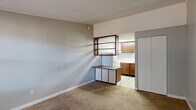 Located at 5520 Collins Road in Jacksonville, Florida, this single-floor studio-style unit offers a practical and efficiently organized layout encompassing a living room, kitchen, bathroom, and closet. The living area features neutral-toned carpet flooring, light gray walls, and a textured ceiling, creating a clean and understated interior. A wall-mounted shelving unit with a built-in desk surface serves as a functional divider between the living space and the kitchen, providing both storage and a dedicated workspace. Natural light enters the living area through a window fitted with horizontal blinds, offering views of surrounding greenery. A wall-mounted HVAC unit ensures year-round climate control. The kitchen is equipped with warm-toned wooden cabinetry, a stainless steel sink with laminate countertops, and light vinyl plank flooring, offering a functional cooking space within a compact footprint. A full-panel closet with bi-fold doors provides convenient storage directly off the living area. The bathroom, accessible from the kitchen corridor, features light vinyl plank flooring, a toilet, a bathtub, and clean white walls. The overall layout is designed to maximize utility within a modest footprint, making it a straightforward and functional living space.