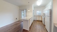 This single-story space is designed with a clean, functional layout highlighted by warm wood-look flooring that flows consistently throughout. The open-concept kitchen serves as a central feature, equipped with white cabinetry, a white electric range, a dishwasher, a refrigerator, and a double sink positioned beneath a window. A peninsula with a light-colored countertop extends into the living area, complemented by a single pendant light overhead, creating a natural divide between the kitchen and living spaces. A secondary window and sliding glass door bring in ample natural light, enhancing the bright, airy atmosphere maintained by white walls throughout. The living area is generously proportioned, with a paneled entry door and multiple wall outlets and registers indicating practical utility. A built-in desk nook with overhead lighting and a countertop surface provides a dedicated workspace. The bathroom features wood-look flooring continuing from the main living areas, a long vanity with an integrated sink, a large mirror, a bathtub, and louvered bifold doors leading to a closet. Overall, the space presents a straightforward, well-lit, and efficiently organized interior.