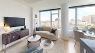 This thoughtfully designed single-floor residence offers a bright, contemporary living experience defined by clean lines, light wood flooring, and an abundance of natural light throughout. The open-concept living area serves as the heart of the home, featuring floor-to-ceiling windows that frame expansive city views. A curved sofa, layered area rug, and curated furnishings create a welcoming yet refined atmosphere, while a structural column adds architectural character to the space.

The kitchen is sleek and functional, outfitted with stainless steel appliances including a gas range and built-in microwave, complemented by gray cabinetry, open shelving, and a subtle tile backsplash. A large window floods the kitchen with natural light and offers striking urban views.

Two well-proportioned bedrooms provide restful retreats. The primary bedroom features large windows with city views, light wood flooring, and access to a dedicated walk-in closet and en-suite bathroom. The en-suite includes a walk-in glass shower, gray vanity cabinetry, and white tile work. The second bedroom is bright and airy, offering generous closet space. The second bathroom features a soaking tub with a tiled surround, a wide vanity, and a large mirror. Throughout, the palette remains consistently neutral and modern, fostering a calm, cohesive ambiance.