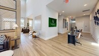Located on Taylor Road in Nashville, TN, this thoughtfully designed community clubhouse offers a generous single-floor layout that blends modern aesthetics with functional living spaces. The main living area makes an immediate impression with soaring vaulted ceilings, exposed wood beam accents, and an eye-catching slatted wood-and-metal room divider that elegantly defines the space. Light wood-tone flooring flows throughout, complemented by crisp white walls that amplify the abundant natural light streaming in from multiple windows. A curated selection of contemporary artwork and greenery, including a striking live moss wall bearing the Sojourn Nashville brand, adds character and warmth to the interior. The combined dining and living room features a stylish kitchen bar area with gray cabinetry, chevron tile backsplash, and a statement chandelier overhead, while large windows dressed in floor-length drapes bathe the space in natural light. The dedicated office is equipped with a built-in workstation, ample cabinetry, floating shelves, and a bold blue shiplap accent wall, offering a productive and well-appointed environment. An additional workstation nook provides supplemental workspace. The covered outdoor balcony extends the living experience with seating for multiple guests and panoramic views of the surrounding Nashville landscape, making it an ideal spot for relaxation or socializing.