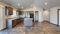 This two-floor residence is thoughtfully laid out to maximize functionality across its living spaces. The main level serves as the heart of the home, featuring an open-concept kitchen and living room area highlighted by vaulted ceilings with recessed lighting that add a sense of openness and height. The kitchen is well-appointed with rich, dark-stained wood cabinetry, granite-style countertops, stainless steel appliances including a range, microwave, and dishwasher, a double-basin sink, and a central island that provides additional workspace and storage. Wood-look tile flooring runs throughout the main living areas, offering both durability and a warm aesthetic. A dedicated pantry closet adds practical storage to the kitchen area. The living room is spacious and bright, with natural light streaming in through windows that offer views of the surrounding exterior. The bedroom is generously sized, featuring soft carpeting, a ceiling fan, a window for natural light, and direct access to the main living area. A dedicated laundry room houses a full-size washer and dryer with overhead shelving. Carpeted stairs with a sleek metal handrail connect the two levels, completing this efficient and modern residence.