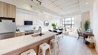 This thoughtfully designed single-floor unit offers an efficient yet stylish living experience across a well-utilized footprint. The open-concept main living area seamlessly integrates the kitchen, dining, and living spaces beneath a distinctive exposed concrete ceiling with track lighting, lending an industrial-modern character to the space. Light hardwood flooring runs throughout, enhancing the bright and airy ambiance.

The kitchen is a standout feature, boasting a long island with a waterfall wood-paneled base, white quartz countertops, a matte black faucet, white upper cabinetry, stainless steel appliances, and a tile backsplash. The dining area comfortably accommodates a full table and chairs, while the living area benefits from expansive floor-to-ceiling windows that flood the space with natural light.

A sleek hallway, accented by a textured concrete accent wall and recessed lighting, connects the main living area to the private quarters. The bedroom is a calm, well-proportioned retreat with high ceilings, light hardwood flooring, and in-unit laundry conveniently tucked within a closet alcove. The bathroom features a clean, all-white aesthetic with subway tile surrounding a full soaking tub and shower combination.