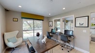 This well-appointed business center is a single-floor space designed to support both collaborative and focused work. The layout is open and functional, featuring light-toned tile flooring throughout that enhances the bright, clean aesthetic of the room. Large windows fitted with horizontal blinds and bold yellow-and-teal valances flood the space with natural light while offering views of the surrounding greenery outdoors.

Along one wall, a dedicated workstation area houses multiple computer desks with sleek, modern monitors and ergonomic chairs, ideal for individual productivity. A large conference-style table with hairpin legs serves as a central gathering point, paired with contemporary black chairs for group meetings or collaborative sessions. Decorative touches, including framed animal-themed artwork, a globe accent piece, and a whimsical ceramic figurine, add character and warmth to the space.

A lounge corner features a sculptural egg chair and a wall-mounted flat-screen television, offering a relaxed setting for informal interactions. A decorative mirror grid panel adds depth and visual interest to the neutral-toned walls. Glass-panel double doors provide direct access to an outdoor area, further connecting the interior to the natural surroundings. Recessed lighting complements the abundant daylight, ensuring the space remains well-lit at all times.