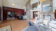 This impressive single-floor space is defined by dramatic architectural details that create a grand yet welcoming atmosphere. The main living area commands immediate attention with its soaring vaulted ceilings, which are accentuated by tall, multi-pane windows that flood the interior with an abundance of natural light. Warm herringbone-patterned hardwood flooring flows throughout, complementing the rich, floor-to-ceiling mahogany wood-paneled walls that lend a refined, sophisticated character to the space.

A prominent natural stone accent wall rises to the full height of the vaulted ceiling, flanking a classic stone fireplace that serves as a stunning focal point in the living area. Comfortable seating is arranged thoughtfully around a glass-top coffee table, creating an inviting lounge setting beneath a statement pendant light fixture. French doors open to an outdoor area, further enhancing the connection between interior and exterior.

A sleek glass-railed staircase with metal framing provides both function and visual interest at the heart of the space. Decorative wall art and a circular mirror add curated touches to the wood-paneled walls.

A corridor with slate tile flooring leads to additional rooms, offering a textural contrast to the main area's hardwood. Throughout, the combination of natural materials, generous ceiling heights, and carefully considered design elements creates a cohesive and distinguished environment.