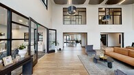 Located in Little Rock, Arkansas, this single-floor community clubhouse is a striking example of contemporary design, offering a generously proportioned and thoughtfully curated gathering space. The grand entrance lobby immediately impresses with its soaring double-height ceilings, dramatic floor-to-ceiling steel-framed windows, and warm light wood flooring that runs throughout the entire space. Clerestory windows and large glass wall panels flood the interior with abundant natural light, creating an airy, open ambiance.

The expansive main living area serves as the heart of the space, anchored by a bold textured accent wall featuring a geometric tile pattern, a linear fireplace, and a wall-mounted television — an ideal focal point for relaxed gatherings. Plush, modern seating arrangements complement the refined aesthetic. Multiple dark wood bar-height islands provide flexible seating and define zones within the open floor plan.

A full kitchen with a striking emerald green tile backsplash, pendant lighting, and bar seating adds functional elegance. The game and recreation wing features a billiards table and casual seating, while a tiled architectural column and water stations add practical amenities. Large sliding glass doors open to outdoor views, seamlessly connecting the interior to the surrounding environment.