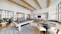 Located at 3200 Meadow Gate Ln in Davenport, FL, this single-floor community living space is a thoughtfully designed gathering area that blends modern aesthetics with inviting functionality. The room is defined by soaring ceilings adorned with prominent exposed natural wood beams, lending warmth and architectural character to the open, airy layout. Underfoot, a striking herringbone-patterned wood floor runs throughout, adding elegance and visual continuity. Floor-to-ceiling steel-framed windows along one wall flood the interior with abundant natural light while offering expansive views of the outdoor amenity area, complete with palm trees and string lighting. The space is organized into distinct activity zones: a lounge area with a plush sectional sofa and a sculptural gold floor lamp; a game zone centered around a light-wood billiard table set atop a layered area rug; and a dining area featuring a round dark-top table with upholstered chairs. At the far end, a well-appointed kitchen station showcases soft blue-gray cabinetry, a long white-topped island lined with woven bar stools, decorative tiled accents, pendant lighting, and a mounted flat-screen TV. A reading nook with shelving is also visible, rounding out the versatile amenities of this well-curated communal space.