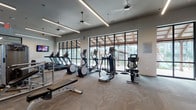 This fitness center offers a comprehensive workout environment designed with both functionality and aesthetics in mind. The space is organized to accommodate a wide range of exercise needs, from cardio training to strength and functional fitness. Large, black-framed floor-to-ceiling windows line one side of the facility, flooding the interior with abundant natural light and providing views of the surrounding outdoor area, creating an open and energizing atmosphere.

The cardio zone features multiple treadmills, elliptical machines, a stationary bike with an interactive display, spin bikes, and a rowing machine, offering diverse options for aerobic training. Overhead, sleek linear pendant lighting and ceiling fans complement the modern, neutral-toned interior, which features herringbone-patterned carpet flooring throughout.

The strength training area is well-appointed with a cable machine, adjustable benches, a rack of free weights, and barbell stations. A functional training wall outfitted with TRX suspension trainers, battle ropes, and resistance bands supports dynamic workouts. Wall-mounted televisions are positioned throughout for entertainment. A built-in cabinetry station with a chalkboard displaying workout suggestions adds a practical, community-focused touch to the space.