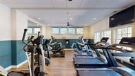Located at 1900 Barclay Blvd in Princeton, NJ, this expansive fitness center offers a comprehensive range of amenities across a generously sized single-floor layout. The facility is divided into two dedicated exercise rooms, a hallway, a changing room, and a multi-purpose court, creating a well-organized environment for a variety of workout routines.

The primary cardio room features a row of modern treadmills positioned in front of large windows that invite natural light and provide pleasant outdoor views of surrounding greenery. Ceiling fans circulate air throughout, and a wall-mounted flat-screen TV adds entertainment for users. A second exercise area is outfitted with strength and resistance machines, free weights, stability balls, and resistance bands, all reflected by full-length mirrors that enhance the sense of space. Light hardwood-style flooring runs throughout the fitness rooms, complementing the two-tone teal and white wall design.

A standout feature of the facility is the full indoor racquetball and basketball court, boasting soaring ceilings, pristine hardwood flooring with red court lines, a regulation basketball hoop, and smooth white walls built to racquetball specifications. The hallway connects all areas seamlessly, with access to changing facilities nearby, making this a fully self-contained athletic destination.