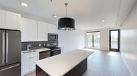 This single-floor, 2-bedroom, 2-bathroom residence showcases a sleek, modern design centered around an open-concept living space. The kitchen serves as a focal point, featuring a large island with a crisp white quartz countertop and dark cabinetry base, creating a striking contrast. White shaker-style upper cabinets line the walls alongside a dark gray subway tile backsplash, complementing the stainless steel appliances, including a built-in microwave and electric range. A bold black drum pendant light hangs above the island, adding a dramatic design element. The kitchen flows seamlessly into the living area, which is bathed in natural light from a large sliding window that offers views to the outdoors. Light wood-tone flooring extends throughout the open living space, enhancing the bright and airy atmosphere. Recessed lighting is installed throughout, ensuring consistent illumination across the entire area. The overall aesthetic is clean and contemporary, combining dark and light contrasts to create a polished, stylish environment ideal for both everyday living and entertaining.