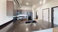 Located at 612 Sixth St in Rapid City, South Dakota, this one-bedroom, one-bathroom unit offers a thoughtfully designed layout with a modern industrial aesthetic. Light hardwood-style flooring flows throughout, complementing crisp white walls and bold dark trim accents.

The kitchen is a standout feature, showcasing rich, medium-toned shaker cabinetry, striking dark stone countertops with a polished finish, and a suite of black appliances including a range, microwave, and refrigerator. An island with a deep undermount sink adds both function and style, while exposed silver ductwork overhead reinforces the space's urban, loft-inspired character.

The bedroom is bright and airy, benefiting from a large picture window that frames views of the Rapid City skyline and surrounding landscape. Exposed ductwork continues here, paired with a ceiling fan and generous natural light. A dedicated walk-in closet off the bedroom provides ample built-in shelving and hanging rods for organized storage.

The bathroom features a clean, neutral palette with light wood flooring, a tub-shower combination with white tile surround, a dark vanity with contrasting hardware, and a matte black shower rod for a cohesive modern touch. A connecting hallway ties all spaces together efficiently, completing this well-planned, contemporary unit.