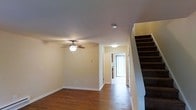 This two-story home offers a straightforward and functional layout designed for comfortable everyday living. The main level features rich hardwood flooring that runs throughout, adding warmth and a classic aesthetic to the space. The living area is equipped with a ceiling fan with integrated lighting, providing both comfort and illumination. Clean, neutral-toned walls create a bright and versatile backdrop, ready to accommodate a variety of interior styles. A hallway extends toward the rear of the main floor, leading to what appears to be a well-lit room or entry point, bathed in natural light filtering through vertical blinds — suggesting a back door or patio access. The staircase, positioned along one side of the main level, features dark-toned treads that contrast neatly with the light walls, providing access to the second floor. The overall layout flows naturally from the living space through the hallway, creating a sense of depth and openness. The home's interior presents a clean, move-in-ready condition with consistent finishes across the visible areas, making it a practical and welcoming two-story residence.