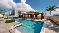 Located at 410 Evernia Street in the heart of West Palm Beach, Florida, this outdoor amenity deck offers a resort-style retreat with stunning open-sky views of the surrounding cityscape. The centerpiece is a generously sized lap pool with clearly marked lanes, ideal for both recreational swimming and fitness, complemented by a separate whirlpool spa tucked within a lush, tropical garden setting. The pool deck features a striking Mediterranean-inspired cabana structure with bold red columns, terracotta-tiled rooflines, and a wooden pergola connecting two pavilion buildings—providing shaded relaxation areas outfitted with blue lounge chairs. The travertine-style deck surface extends the full length of the pool, offering ample space for sunbathing and leisure. Mature palm trees, vibrant tropical greenery, and manicured hedges frame the perimeter, creating a private, garden-like atmosphere. Panoramic views of the West Palm Beach skyline and neighboring high-rises add an urban sophistication to the otherwise serene outdoor environment. The space is well-appointed with safety equipment and signage, reflecting a thoughtfully maintained community amenity designed for both relaxation and active outdoor living under the sunny South Florida sky.