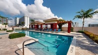 Located at 410 Evernia Street in the heart of West Palm Beach, Florida, this outdoor amenity deck offers a resort-style retreat with stunning open-sky views of the surrounding cityscape. The centerpiece is a generously sized lap pool with clearly marked lanes, ideal for both recreational swimming and fitness, complemented by a separate whirlpool spa tucked within a lush, tropical garden setting. The pool deck features a striking Mediterranean-inspired cabana structure with bold red columns, terracotta-tiled rooflines, and a wooden pergola connecting two pavilion buildings—providing shaded relaxation areas outfitted with blue lounge chairs. The travertine-style deck surface extends the full length of the pool, offering ample space for sunbathing and leisure. Mature palm trees, vibrant tropical greenery, and manicured hedges frame the perimeter, creating a private, garden-like atmosphere. Panoramic views of the West Palm Beach skyline and neighboring high-rises add an urban sophistication to the otherwise serene outdoor environment. The space is well-appointed with safety equipment and signage, reflecting a thoughtfully maintained community amenity designed for both relaxation and active outdoor living under the sunny South Florida sky.