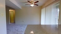 This single-floor residence offers a comfortable and functional layout designed for everyday living. The main living area is open and inviting, featuring plush wall-to-wall carpeting that adds warmth underfoot. A classic five-blade ceiling fan with an integrated light fixture is centrally mounted, providing both air circulation and ambient lighting throughout the space. Smooth, neutral-toned walls create a clean, versatile backdrop suited to a variety of interior styles. Natural light filters generously into the room through large floor-to-ceiling vertical blinds, which offer both privacy and light control while enhancing the sense of openness. A prominent granite-topped counter is visible in the foreground, its speckled surface lending a touch of durability and style, likely serving as a dividing element between the living and dining or kitchen areas. The layout flows naturally between spaces, making the home feel cohesive and well-proportioned. Overall, the residence presents a straightforward, well-maintained interior with practical finishes and a bright, airy ambiance that makes it a comfortable place to call home.
