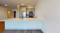 This single-floor residence at 444 Franklin Street in Framingham, Massachusetts offers a clean, contemporary design with a thoughtful layout. The open-concept living and kitchen area is anchored by warm hardwood-style flooring that extends throughout the main living space, creating a cohesive and inviting atmosphere. The kitchen is well-equipped with light wood cabinetry featuring matte black hardware, a blue subway tile backsplash, stainless steel appliances including a refrigerator, microwave, and range, and a generous white quartz peninsula with an undermount sink. Stylish pendant lights with gold accents hang above the peninsula, adding a refined touch. The living area is spacious and bright, with recessed lighting and easy access to the bedroom wing. Bedrooms feature plush carpeting and large windows fitted with horizontal blinds, allowing natural light while offering views of the surrounding neighborhood. At least one bedroom includes a walk-in closet with built-in shelving and hanging rods, providing ample storage. The bathroom is finished with large-format tile flooring, a light wood vanity with a white countertop and integrated sink, a mirror, and a tiled shower/tub combination, rounding out this modern and functional living space.