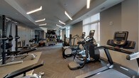 Located at 1100 2nd Place SE in Washington DC, this modern fitness center is a well-appointed, single-floor facility designed to support a full range of workout needs. The space features a clean, contemporary aesthetic with neutral gray tones, herringbone-patterned carpet flooring, and dedicated rubber matting zones for weightlifting and functional training areas.

Overhead, rows of sleek linear pendant lighting illuminate the expansive floor plan, complemented by natural light streaming through large windows fitted with roller shades. Full-length mirrors line sections of the walls, enhancing the sense of openness and allowing users to monitor their form.

The cardio zone is equipped with multiple elliptical machines, stationary bikes, and treadmills, all featuring digital displays. The strength training area includes a robust dumbbell rack with a wide range of weights, a power rack, and various benches for free weight exercises. A functional training station rounds out the space with medicine balls, kettlebells, resistance equipment, and TRX suspension trainers neatly organized on a multi-tier rack.

A glass-walled studio area adjacent to the main floor offers a dedicated space for group classes or stretching, with light wood-look tile flooring. Dual water fountains with bottle-filling stations are conveniently located near the studio entrance, adding a practical and user-friendly touch to this comprehensive fitness facility.