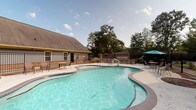 This community pool and clubhouse facility offers a well-rounded outdoor and indoor experience. The centerpiece is a generously sized freeform swimming pool with clear, turquoise water, surrounded by a wide concrete deck furnished with lounge chairs, shaded seating areas with umbrellas, and tables for outdoor dining. A black iron perimeter fence encloses the pool area, with mature trees providing a natural backdrop and added privacy.

The adjoining clubhouse is designed with soaring vaulted ceilings that lend the interior a bright and airy character. Rich hardwood-style flooring flows throughout, complementing the warm neutral tones of the walls. The main gathering room is generously proportioned, featuring multiple ceiling fans, large windows that invite natural light, and a tall arched window that draws the eye upward. This room accommodates both lounge-style seating with plush leather sofas and multiple round dining tables with high-back chairs, making it suitable for both casual relaxation and group gatherings.

A pass-through bar connects to a compact kitchen area, complete with dark cabinetry and bar stool seating — ideal for entertaining. Additional amenities include restrooms and a dedicated office or service window, ensuring the space functions efficiently for residents and guests alike.