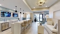 This beautifully appointed clubhouse delivers a sophisticated and welcoming atmosphere across a single, open floor. The space is anchored by a long, white quartz bar-height counter lined with woven rattan-backed barstools, creating a social focal point ideal for gathering and relaxing. Overhead, a striking starburst chandelier with gold metallic arms serves as a dramatic design centerpiece, complemented by sleek black pendant lights that add a modern contrast to the room's light, neutral palette. The flooring features large-format light-toned tiles that enhance the sense of openness and cleanliness throughout. A wall-mounted flat-screen television is positioned above a built-in console, providing both function and entertainment. The lounge area is furnished with plush upholstered seating, including velvet-toned accent chairs and a clean-lined sofa, offering comfortable spots for conversation or leisure. Decorative wallpaper with a subtle mosaic pattern adds a touch of texture and visual interest. Floor-to-ceiling glass doors and oversized windows line the far wall, flooding the interior with natural light and offering views of the surrounding outdoor area. The overall ambiance is bright, modern, and refined, making this clubhouse an inviting retreat for residents and guests alike.
