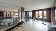 This modern fitness center is thoughtfully designed to support a wide variety of training disciplines within a single, open-plan space. The layout is anchored by expansive hardwood-style flooring that provides a clean, durable surface throughout. Deep plum-toned walls lend a sleek, contemporary aesthetic, contrasted by crisp white ceilings with recessed lighting that ensures the space is evenly and brightly lit.

A full wall of floor-to-ceiling mirrors dominates one side of the room, visually expanding the space and supporting form-focused training. Mounted opposite is a large flat-screen display, ideal for guided workouts or virtual classes. The functional training zone is well-stocked with equipment including stability balls, balance boards, medicine ball towers, colorful kettlebells, TRX suspension systems, plyo boxes, and resistance tools.

A dedicated cycling area houses multiple stationary bikes positioned in front of tall, vertical windows that frame sweeping elevated views of the surrounding cityscape and greenery — an inspiring backdrop for cardio sessions. Additional large windows line multiple walls throughout the space, flooding the interior with abundant natural light.

Glass partitions and metal railing details add an architectural refinement, while the open floor plan allows for flexible class arrangements or individual training sessions. The overall environment is energetic yet polished, designed for serious fitness use.