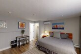 Located at 473 Main St in Hyannis, MA, this efficiently designed single-floor suite offers a smartly organized layout that maximizes every inch of its space. The main room serves as both a bedroom and living area, featuring warm hardwood-style flooring, a textured ceiling, and wainscoting along the lower walls that adds a classic coastal charm. A wall-mounted mini-split unit provides dedicated climate control, while a large flat-screen TV mounted above a built-in workspace with white cabinetry, a mini-refrigerator, and a microwave creates a functional kitchenette-style nook. Nautical-themed artwork and colorful wall pieces contribute to the room's coastal New England character. A window dressed with drape panels allows natural light to filter in. The bedroom area accommodates a queen-sized bed with a bedside table and lamp, alongside a small dining table with stools. A dedicated closet offers additional storage. The private bathroom features classic black-and-white diamond-patterned tile flooring, a white vanity with sink, a shower with curtain, and a bright window that brings in natural light. This suite is ideal for those seeking a practical and charming retreat in the heart of Hyannis.