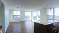 This single-floor space is defined by its clean, modern design and an abundance of natural light. The open-concept layout creates a seamless flow between the living and kitchen areas, making the space feel expansive and airy. Large floor-to-ceiling windows wrap around the corner of the living area, flooding the interior with daylight and offering sweeping views of the surrounding cityscape and greenery beyond. The windows are fitted with horizontal blinds, allowing for flexible light control throughout the day. Warm hardwood flooring runs throughout the living space, adding a rich, natural contrast to the crisp white walls and ceilings. Recessed lighting is strategically placed throughout the ceiling, complementing the natural light and providing a clean, uncluttered overhead aesthetic. A contemporary pendant light fixture hangs near the kitchen area, adding a refined accent. The kitchen features a substantial dark-toned peninsula with a bright white countertop, offering both functional workspace and a striking visual contrast. The overall ambiance is bright, minimalist, and sophisticated, making it a versatile and welcoming environment suited to a variety of uses.