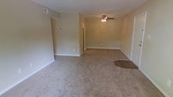 Located at 301 Village Dr in Jacksonville, North Carolina, this single-floor residence offers a straightforward and functional layout designed for comfortable everyday living. The main living area is generously proportioned, featuring wall-to-wall carpeting in a neutral beige tone that creates a warm and inviting atmosphere throughout the space. Smooth, light-colored walls provide a clean backdrop, complemented by white baseboards that frame each room with a classic, tidy finish. A ceiling fan with an integrated light fixture is centrally mounted overhead, offering both air circulation and ambient lighting to the living area. The open layout allows for flexible furniture arrangement, making it easy to define distinct living and dining zones within the same shared space. A transitional area with a patch of hardwood-style flooring near an entryway adds a subtle contrast to the carpeted surroundings. Doorways lead to additional rooms, including what appears to be a kitchen visible in the background, continuing the home's consistent neutral color palette. The overall ambiance is clean, understated, and move-in ready, suited for those seeking a practical and low-maintenance living environment in the Jacksonville area.