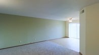 This single-floor space presents a straightforward and functional layout designed for comfortable everyday living. The main living area is characterized by soft green walls that lend a calm, neutral ambiance to the room. Underfoot, plush wall-to-wall carpeting adds warmth and comfort, while a section of hard flooring near the entrance or transition area provides a practical contrast. A ceiling fan with a light fixture is centrally positioned overhead, ensuring year-round comfort and adequate illumination. A large window dressed with vertical blinds allows natural light to filter into the space, brightening the interior and creating an airy feel. The textured ceiling adds a subtle architectural detail consistent throughout the room. Clean baseboard trim lines the perimeter of the walls, contributing to a tidy, well-maintained appearance. The open layout of the living area offers flexibility for a variety of furniture arrangements, making it an adaptable space suited to different lifestyle needs. Overall, the space conveys a simple, move-in-ready quality with a quiet, understated aesthetic.