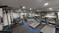 This fitness room at 143 Stoneridge Dr in Columbia, South Carolina, offers a efficiently organized exercise space designed to maximize functionality within its compact footprint. The room features neutral gray walls paired with recessed lighting that provides consistent, even illumination throughout the space, creating a clean and focused workout environment.

A standout design element is the large wall-to-wall mirror installation along one side of the room, which visually expands the space and allows users to monitor their form during exercise. The flooring consists of modular carpet tiles arranged in a bold chevron pattern, combining gray and blue tones to add visual interest and define the workout area.

The room is equipped with multiple cardio machines, including treadmills and an elliptical trainer, as well as a multi-function strength training machine, offering a well-rounded selection of fitness options. A sanitization station and fire safety equipment are also present near the entrance, reflecting attention to health and safety standards.

Access is provided through a glass-paneled door, and ventilation is maintained via ceiling vents. The overall ambiance is modern and purposeful, delivering a streamlined fitness experience suitable for a variety of workout routines.