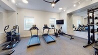 Located at 2110 Pickens St. in Columbia, SC, this fitness center is a thoughtfully designed, single-floor workout facility that combines functionality with a clean, modern aesthetic. The space is defined by crisp white walls, grey rubber flooring, and a combination of track lighting and ceiling-mounted fixtures that keep the environment well-lit throughout.

The cardio zone is generously outfitted with multiple TRUE treadmills, an elliptical trainer, a stationary air bike, and a rowing machine, providing a full range of cardiovascular training options. Natural light filters in through several windows adorned with decorative ironwork grilles, lending the space a bright and welcoming atmosphere. A wall-mounted flat-screen TV and a large ceiling fan add comfort for longer workout sessions.

A striking floor-to-ceiling glass partition with a bold black steel frame visually divides the fitness area from an adjacent hardwood-floored room, adding an architectural element that enhances the sense of openness. The strength training section is equipped with a full power rack with barbells and weight plates, a dumbbell rack with a comprehensive set of free weights, and an adjustable workout bench. A wall of mirrors further amplifies the sense of space and supports proper form during training. The overall design balances practicality with a polished, professional feel.