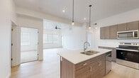 This well-appointed single-floor residence offers two bedrooms and two bathrooms within a thoughtfully designed layout. The open-concept kitchen serves as a central feature, showcasing a large island with a double-basin sink, sleek white quartz countertops, wood-toned cabinetry, and a full suite of stainless steel appliances including a range, microwave, and refrigerator. Pendant lighting above the island adds a modern touch, while light hardwood-style flooring extends throughout the main living areas.

The kitchen flows seamlessly into the living room, which is brightened by natural light from large windows fitted with blinds and accented by a ceiling fan overhead. The two bedrooms feature neutral carpet flooring and ample natural light. The primary bedroom connects to a well-sized walk-in closet with built-in shelving and hanging rods for organized storage.

Both bathrooms are finished in a contemporary style, with the primary bathroom featuring a dual-sink vanity with wood-toned cabinetry, a large mirror with modern lighting fixtures, and light wood flooring. A dedicated laundry area houses a full-sized side-by-side washer and dryer. The overall aesthetic is clean, bright, and modern throughout.