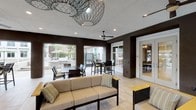 This covered outdoor amenity space seamlessly blends indoor comfort with open-air living. The area is designed with a sophisticated palette, featuring deep espresso-toned structural columns contrasted against warm natural stone masonry walls that add texture and visual interest throughout. Large open bays frame unobstructed views of the pool deck and palm tree-lined surroundings, flooding the space with natural light and a sense of expansiveness.

The lounge area anchors one section of the space, furnished with a generously sized neutral-toned sofa and coordinating accent chairs with wooden armrests and perforated metal panels. A striking sculptural wire pendant light serves as a dramatic focal point overhead, complementing the recessed lighting embedded in the smooth white ceiling. Ceiling fans are strategically positioned throughout to ensure comfortable airflow.

A dedicated dining zone offers counter-height tables and dark-framed chairs suitable for casual gatherings. Light-toned large-format tile flooring runs continuously throughout, enhancing the airy, clean aesthetic. A fireplace integrated into the stone wall adds a functional and architectural element to the space. Horizontal dark metal privacy screens provide partial enclosure while maintaining the open, resort-inspired atmosphere. The overall ambiance is modern, polished, and well-suited for social gatherings and relaxed outdoor living.
