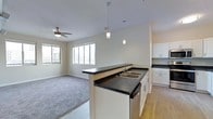 This single-floor space is designed around an open-concept layout that seamlessly connects the kitchen and living area. The kitchen is anchored by a peninsula with dark countertops, offering a double sink and additional prep space. White cabinetry lines the surrounding walls, providing generous storage, while pendant lighting hangs above the peninsula, adding focused illumination and a modern aesthetic touch. The kitchen transitions into the living area, where light hardwood-style flooring gives way to plush carpeting, delineating the two zones without the use of walls. The living area is bright and airy, benefiting from multiple windows that draw in natural light and offer views of the surrounding exterior. A ceiling fan is centrally positioned in the living space, contributing to both comfort and airflow. The neutral wall tones throughout create a clean, versatile backdrop suited to a variety of furnishing styles. The overall ambiance is practical and modern, with an emphasis on open sightlines and functional design that makes the space feel connected and welcoming.