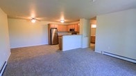 This single-floor residence offers a thoughtfully designed open-concept layout that seamlessly connects the kitchen and living areas into one cohesive, functional space. The kitchen is well-appointed with light maple cabinetry, granite-style countertops, and stainless steel appliances including a full-size refrigerator and microwave. A peninsula-style breakfast bar extends from the kitchen, providing additional counter space and a natural dividing point between the cooking and living areas. The living area is finished with neutral-toned wall-to-wall carpeting that adds warmth and comfort underfoot. A ceiling fan with integrated lighting serves the main living space, ensuring both comfort and illumination. The layout includes a secondary area visible beyond the kitchen, hinted at by a glimpse of additional cabinetry and natural light, suggesting a bathroom or utility space. The overall color palette is warm and neutral, with light walls and wood-toned finishes creating a welcoming ambiance throughout. The open floor plan maximizes the sense of space and allows for flexible furniture arrangement, making it a practical and comfortable living environment.