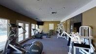 This single-floor fitness center offers a comprehensive workout environment designed to meet a variety of exercise needs. The space is outfitted with a solid selection of cardio equipment, including a Precor elliptical and a stationary bike, providing residents or members with quality machines for endurance training. A dedicated strength training area features a multi-function cable machine, a free weight rack with dumbbells, and additional resistance equipment, giving users the tools for a full-body workout routine.

The room is finished with durable, low-pile carpet flooring in a neutral tone, complementing the warm olive and charcoal color scheme along the walls. Recessed lighting throughout the textured ceiling ensures the space remains well-lit during any hour. A wall-mounted flat-screen television adds entertainment value for those looking to stay engaged during longer cardio sessions.

Large glass-paned doors along one wall allow natural light to filter into the space while offering a view of the outdoors, contributing to an open and energizing atmosphere. A whiteboard and informational display are mounted on the wall, providing scheduling or instructional resources. Floor-to-ceiling mirrored panels on one side enhance the sense of spaciousness while supporting proper form during strength exercises.