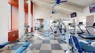 Located at 5700 Market St in Prescott Valley, Arizona, this fitness center offers a single, open exercise room designed to support a comprehensive workout experience. The space is defined by its striking architectural character, most notably a soaring vaulted ceiling with white-paneled slats and large blue ceiling fans that promote airflow throughout the room. Bold red-and-white striped accent walls and vibrant mosaic-style wall art give the space an energetic, motivating atmosphere.

The flooring features a classic checkerboard pattern in gray and tan rubber tile, providing both durability and visual appeal. Large windows along multiple walls flood the room with abundant natural light and offer views of the surrounding Prescott Valley landscape, enhancing the open and airy feel of the space.

The exercise room is well-stocked with a variety of equipment. Cardio offerings include treadmills, stationary spin bikes, and an elliptical machine, each equipped with individual display screens. Strength training options include a cable crossover machine, weight benches, selectorized resistance machines, and a dumbbell rack. Medicine balls and additional accessories are also available. Multiple wall-mounted flat-screen televisions are positioned throughout the room for entertainment during workouts. Full-length mirrors along one wall further expand the sense of space while allowing users to monitor their form.