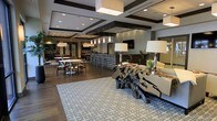 This elegantly designed single-floor space serves as a refined communal gathering area, blending modern sophistication with inviting warmth. The open-concept layout seamlessly integrates multiple functional zones, creating a fluid and versatile environment.

The space is anchored by rich hardwood flooring that runs throughout, complemented by a coffered ceiling with bold dark beams and recessed square light panels that add architectural depth overhead. Pendant lighting in clusters and large drum-shade floor lamps provide layered illumination, contributing to the space's upscale ambiance.

A central bar area with textured, tile-faced paneling serves as a focal point, furnished with sleek bar stools and backed by soft, tufted seating. A distinctive live-edge driftwood coffee table adds an organic, artistic element to the lounge seating area, which is anchored by a large geometric patterned area rug.

A separate dining zone features bistro-style tables and contemporary mixed-material chairs, ideal for casual gatherings. In the background, a kitchenette or café counter area is visible, suggesting additional amenity functions. Lush tropical greenery punctuates the space with natural color, enhancing the overall welcoming atmosphere. The result is a polished, multi-use lounge environment designed for both socializing and relaxation.