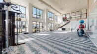This two-story fitness center is a well-appointed facility designed to support a comprehensive range of workout routines. The main floor is an expansive, open exercise area finished with a patterned blue-and-white carpet and soaring double-height ceilings. Large, floor-to-ceiling windows line the perimeter, flooding the space with natural light and providing views of an outdoor pool and surrounding grounds. A ceiling fan overhead and exposed ductwork contribute to the space's modern industrial aesthetic.

The main floor is equipped with a full suite of cardio machines, including treadmills, ellipticals, stair climbers, and rowing machines, as well as a comprehensive selection of strength-training equipment such as cable machines, weight benches, a punching bag, and free weights. A wall-mounted TV, mirrored wall, water fountain, and storage cubbies add functional convenience. A teal tile accent wall adds a distinctive design element to the space.

A staircase with a contemporary metal railing leads to the second floor, which houses a dedicated group fitness and yoga studio. This upper-level room features a smooth, light-toned floor, a wall-mounted barre, large windows, and a TV screen. It is stocked with medicine balls, kettlebells, resistance bands, and other functional training accessories, making it ideal for group classes or individual training sessions.