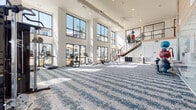 This two-story fitness center is a well-appointed facility designed to support a comprehensive range of workout routines. The main floor is an expansive, open exercise area finished with a patterned blue-and-white carpet and soaring double-height ceilings. Large, floor-to-ceiling windows line the perimeter, flooding the space with natural light and providing views of an outdoor pool and surrounding grounds. A ceiling fan overhead and exposed ductwork contribute to the space's modern industrial aesthetic.

The main floor is equipped with a full suite of cardio machines, including treadmills, ellipticals, stair climbers, and rowing machines, as well as a comprehensive selection of strength-training equipment such as cable machines, weight benches, a punching bag, and free weights. A wall-mounted TV, mirrored wall, water fountain, and storage cubbies add functional convenience. A teal tile accent wall adds a distinctive design element to the space.

A staircase with a contemporary metal railing leads to the second floor, which houses a dedicated group fitness and yoga studio. This upper-level room features a smooth, light-toned floor, a wall-mounted barre, large windows, and a TV screen. It is stocked with medicine balls, kettlebells, resistance bands, and other functional training accessories, making it ideal for group classes or individual training sessions.