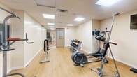 Located at 120 Shelborne Ave in Toronto's Glencairn neighbourhood, this dedicated exercise room offers a well-equipped fitness environment within a compact, efficiently organized footprint. The space features light wood laminate flooring throughout, which contributes to a clean and energetic atmosphere, while a drop tile ceiling with recessed panel lighting ensures the room is consistently and evenly lit.

The room is outfitted with a diverse selection of fitness equipment suited to both cardio and strength training. Cardio options include a SportsArt treadmill, an elliptical trainer, and a stationary bike, providing variety for aerobic workouts. A multi-tiered dumbbell rack stocked with a full range of free weights is prominently positioned, alongside a flat/incline weight bench and a cable resistance machine for strength-focused training. Medicine balls and additional accessories round out the equipment selection. A wall-mounted television is also present, enhancing the workout experience.

A floor-to-ceiling mirror along one wall visually expands the space and assists users in monitoring their form during exercise. The neutral beige walls and white baseboards create a clean, functional backdrop. HVAC vents are integrated into the ceiling for climate control, and a secured exit door with a window provides safe egress. Overall, this fitness room delivers a practical and well-appointed space for a comprehensive workout routine.