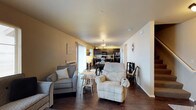 This two-floor space offers a comfortable and functional layout anchored by an open-concept main living area. The living room is furnished with plush seating and flows seamlessly into the dining and kitchen areas, creating a connected and sociable environment. Dark hardwood-style flooring runs throughout the main level, providing a grounding contrast to the warm beige and white walls.

The kitchen is equipped with dark cabinetry, black appliances including a refrigerator and microwave, and is illuminated by a pendant chandelier that adds a subtle decorative touch above the dining area. The dining space accommodates a table with chairs, positioned conveniently between the kitchen and living room.

Natural light enters through a sliding glass door or large window on the far wall of the living room, brightening the space and offering a connection to the outdoors. Neutral wall tones and consistent flooring throughout the main level contribute to a cohesive and inviting ambiance. The second floor provides additional living space, expanding the overall footprint of this well-organized home.
