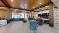 This expansive single-floor lobby and common area blends contemporary design with warm, natural materials to create an inviting and functional gathering space. The main living area immediately draws the eye with its striking natural wood plank ceiling, accented by recessed lighting and bold dark structural columns that define the space without enclosing it. Large-format tile flooring flows seamlessly throughout, lending a clean, polished foundation to the open layout.

At the heart of the living area, a curated seating arrangement of upholstered armchairs in muted blue and gray tones is anchored by a patterned area rug, encouraging conversation and relaxation. Adjacent to this, a built-in counter with bar-height seating sits against a dramatic stone accent wall, offering a casual workspace or social setting. A full-height stacked stone fireplace serves as a focal point in a secondary lounge, framed by a statement ring chandelier and accompanied by a plush sofa and armchairs for comfortable gathering.

A dedicated reading nook features floor-to-ceiling built-in shelving filled with books and décor, paired with gold-accented lounge chairs and large windows that invite natural light and outdoor views. The hallway connects seamlessly to the main areas, featuring a mailbox station and steel-framed glass partitions that add an industrial-modern touch while maintaining an open, airy feel throughout.