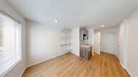 Located at 16210 NE 80th St in Redmond, WA, this efficiently designed studio unit offers a thoughtfully organized single-floor layout that maximizes every square foot of available space. The kitchenette is a standout feature, showcasing sleek gray wood cabinetry, a marble-style backsplash and countertop, an integrated sink, and a built-in microwave. Adjustable open wire shelving adjacent to the kitchen provides practical additional storage. The main living area is bright and open, finished with warm hardwood-style flooring that runs throughout the unit, creating a cohesive and clean aesthetic. Crisp white walls and recessed lighting contribute to a light, airy ambiance. A window fitted with horizontal blinds allows natural light to filter in while offering views of surrounding greenery, adding a pleasant outdoor connection to the interior. The bathroom is clean and functional, featuring a shower/tub combination, a toilet, chrome towel bar, and a vanity, all finished in a simple, modern style consistent with the rest of the unit. The overall design is minimalist and move-in ready, making efficient use of the compact footprint while maintaining a comfortable and practical living environment.