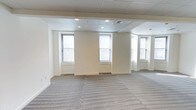 This single-floor commercial space offers a clean, versatile layout well-suited for a variety of office or professional uses. The open floor plan is finished with patterned, neutral-toned carpet tiles that lend a polished yet practical feel underfoot. Walls are painted in a soft, warm white, creating a bright and welcoming atmosphere throughout.

The drop ceiling is fitted with a grid of acoustic tiles and evenly spaced recessed lighting, ensuring consistent illumination across the space. Multiple windows, framed with classic white trim and equipped with blinds, line the perimeter walls and invite ample natural light, enhancing the overall sense of openness. A curved bay window section at one end adds a distinctive architectural touch, breaking up the standard rectangular footprint and offering additional light exposure.

Perimeter baseboard heating units are visible beneath the windows, providing practical climate control. The space transitions smoothly between areas, with no visible interior partitions, making it highly adaptable for open-plan office configurations or customized build-outs. Overall, the space presents a neutral, move-in-ready canvas with classic architectural details, functional infrastructure, and a bright, professional ambiance ideal for a range of commercial occupants.